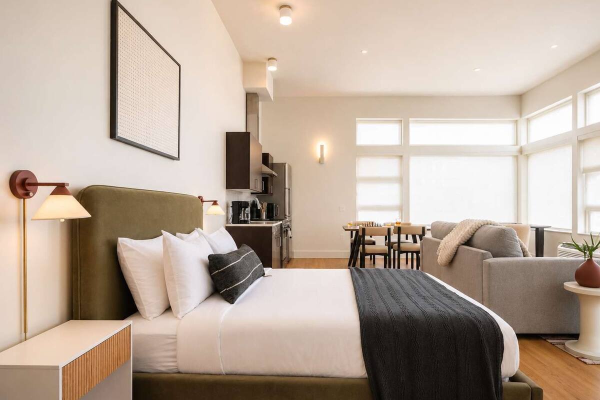 A modern hotel room with a bed, a desk, a sofa, and a kitchenette.