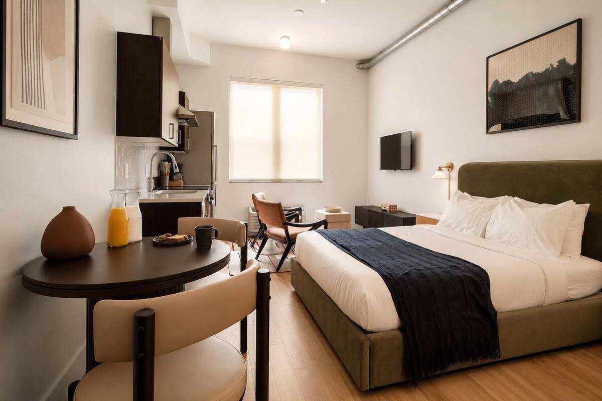 A modern hotel room with a bed, a desk, a chair, a small kitchen area, and a TV.