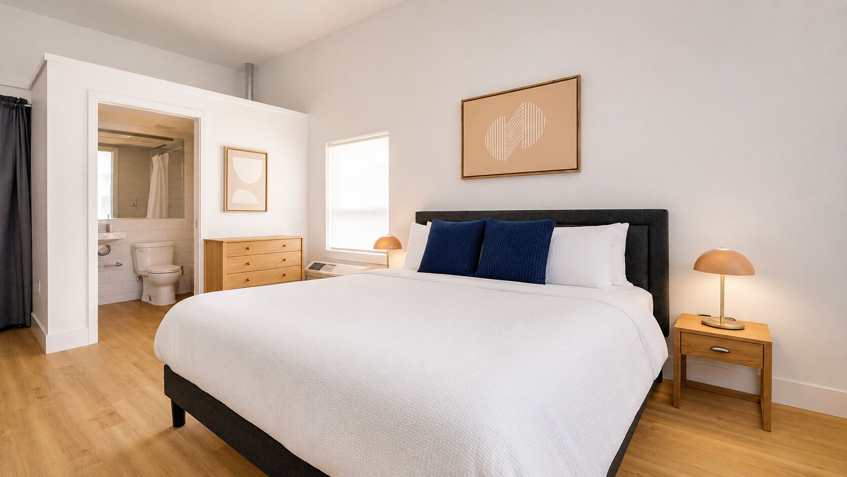 Un dormitorio con cama, mesitas de noche, una ventana, una puerta que da a un baño y un cuadro en la pared.