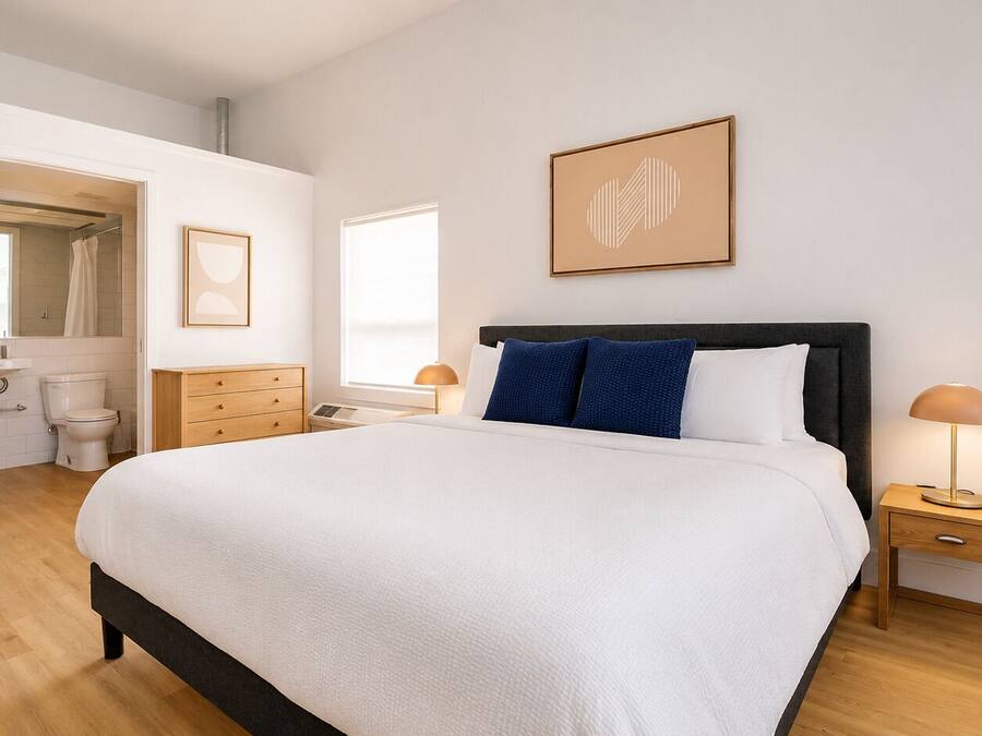 Un dormitorio con cama, mesitas de noche, una ventana, una puerta que da a un baño y un cuadro en la pared.