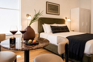 A hotel room with a bed, bedside lamps, a table with a candle, wine glasses, and a vase with a plant.