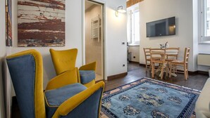 Ein Wohnzimmer mit gelben und blauen Sesseln, einem Essbereich mit Holzsesseln, einem an der Wand montierten Fernseher und einem gemusterten Teppich.