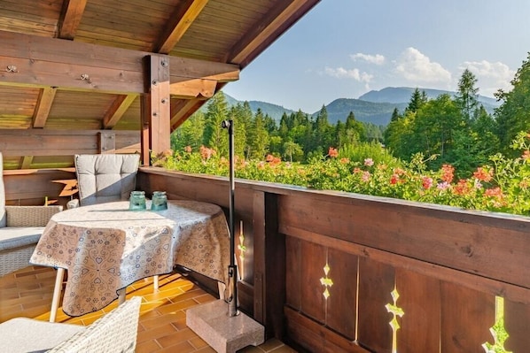 Eine Holzterrasse mit Tisch und Sitzgelegenheiten, mit Blick auf eine Berglandschaft mit Bäumen und Blumen.