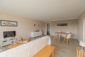 Ein modernes Wohnzimmer mit einem Flachbildfernseher, einem weißen Sofa, einem Esstisch mit Stühlen und einer kleinen weißen Kommode.