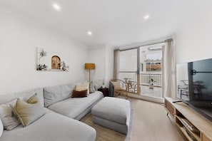 Ein Wohnzimmer mit einer Couch, einem Fernseher, einer Lampe und einem Balkon mit Gartenmöbeln.