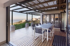 Un espace extérieur aménagé pour les repas, avec une table et des chaises, une pergola et une vue sur la mer et les bâtiments.