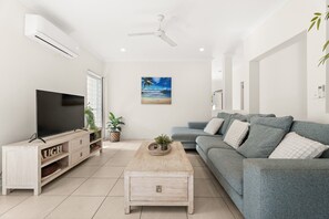 Una sala de estar con televisor de pantalla plana, un sofá gris, una mesa de centro de madera y un ventilador de techo.