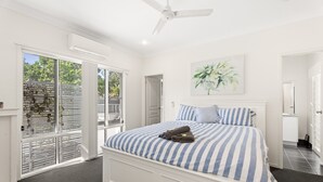 Une chambre avec un grand lit, un ventilateur de plafond, une fenêtre avec des stores et un tableau au mur.