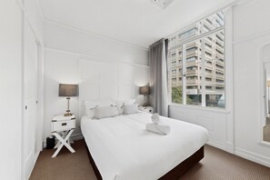 Ein Hotelzimmer mit einem großen Bett, einem Nachttisch mit Lampe, einem Fenster mit Blick auf Gebäude und einer Tür.