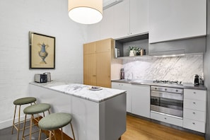 Eine moderne K üche mit Marmorarbeitsplatte, Holzstühlen, einem eingebauten Backofen und einem gerahmten Kunstwerk an der Wand.