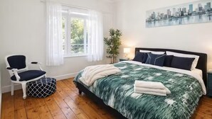 Ett sovrum med en säng, en stol, en växt och en målning av en stadsbild på väggen.