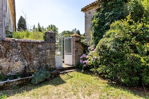 Un muro de piedra, un portón blanco y abundante vegetación.