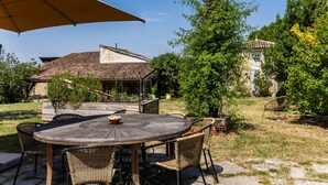 A round wooden table with wicker chairs, a beige umbrella, and a view of a traditional building with a tiled roof. - Guest Room “Famille Cabernet Ch Cajus” with Shared Pool, Terrace, and Wi-Fi (Saint-Germain-du-Puch)