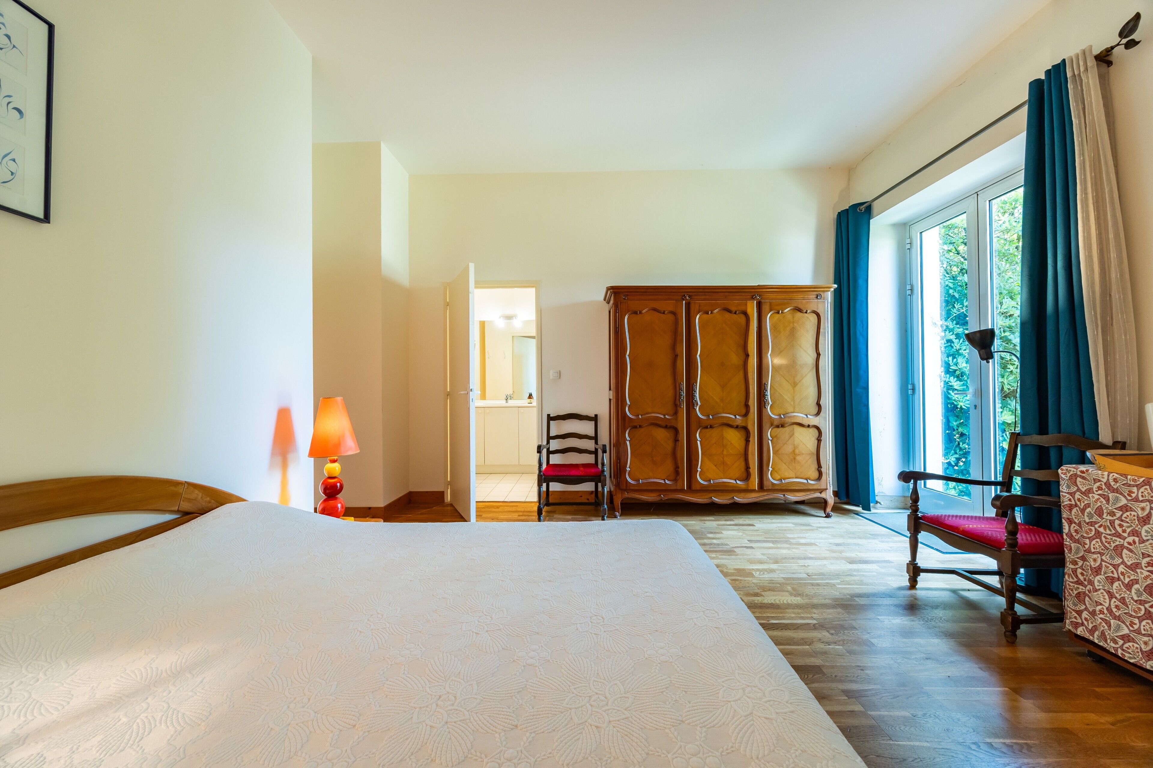 A hotel room with a bed, a wardrobe, a chair, a table, and a view of the outdoors.