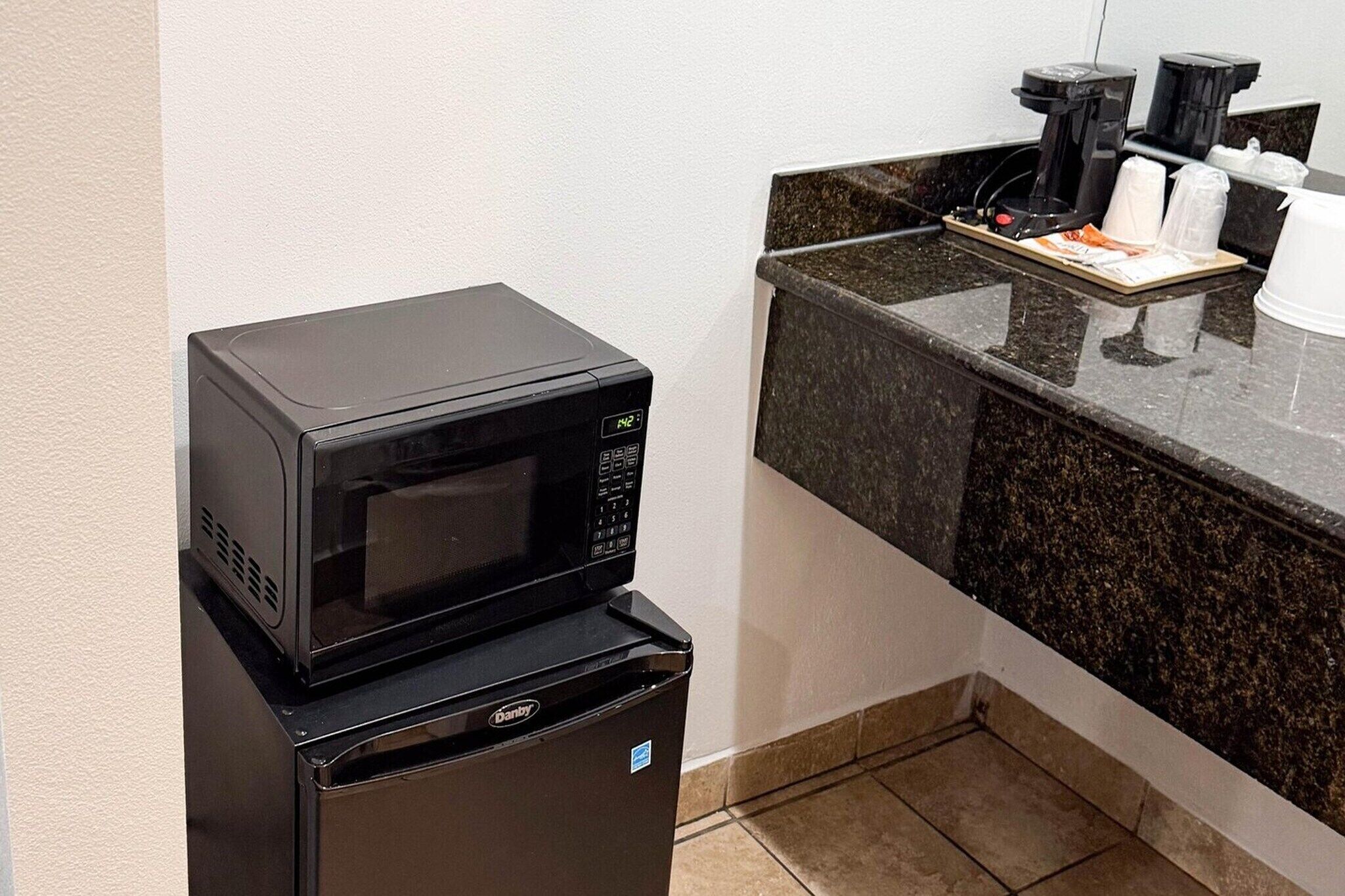 Un petit coin cuisine comprenant un micro-ondes posé sur un réfrigérateur, une machine à café et un plateau avec des gobelets jetables.