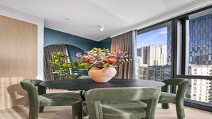 A modern room with a round table, green upholstered chairs, a vase with flowers, and a city view through large windows.