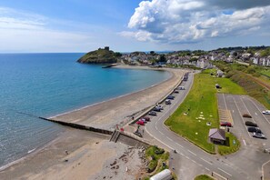 A coastal road with a beach, a parking lot, and a hill with a structure. - Finest Retreats | Awel Y Mor (Criccieth)