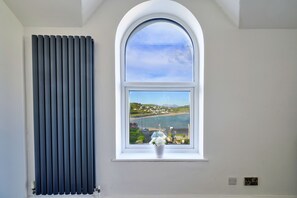 A room with a radiator, a window with a view of a coastal area, and a vase with flowers on the windowsill. - Finest Retreats | Gwynle (Criccieth)