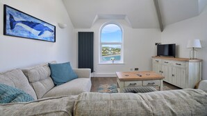 A room with a sofa, a table, a TV, a cabinet, a window with a view, and a painting of a whale. - Finest Retreats | Gwynle (Criccieth)