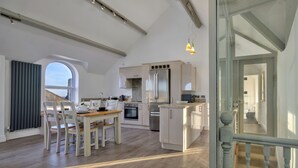 A modern kitchen with white cabinetry, a central island, and a dining area with a table and chairs. - Finest Retreats | Gwynle (Criccieth)