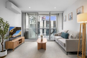 Una sala de estar moderna con un sofá, una mesa de centro, un televisor sobre un mueble de madera, una planta en maceta y un balcón con vistas a la ciudad.