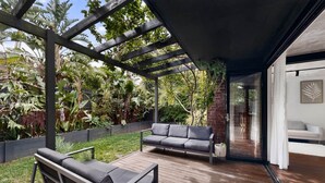 Een houten terras met een pergola, twee grijze banken en een glazen deur die naar een wit interieur leidt.