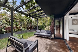 A wooden deck with a pergola, two grey sofas, and a glass door leading to a white interior. - Sunlit 3-Bed Coastal Stay with Private Deck (Cronulla)
