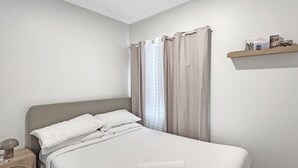 En snyggt bäddad säng med vita sängkläder, ett sängbord med en lampa och en hylla med böcker och dekorativa föremål.