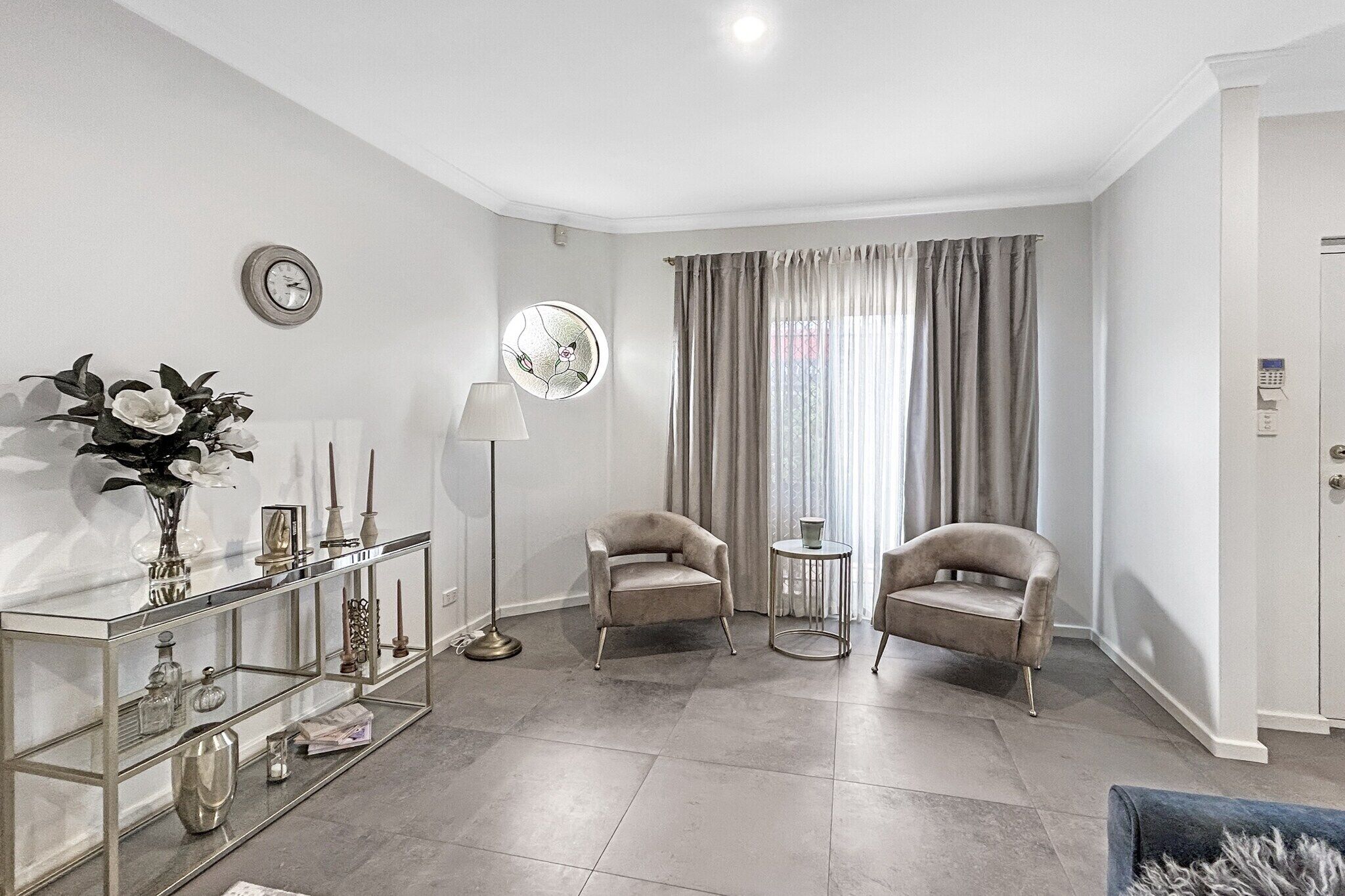A modern living room with a glass console table, two armchairs, a round side table, a floor lamp, and a window with curtains.