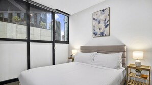 Una habitación de hotel con una cama grande, dos mesitas de noche con lámparas, un ventanal con vistas a edificios y una obra de arte floral en la pared.
