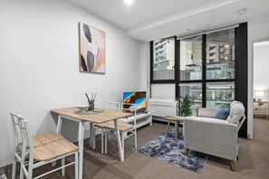 A modern living room with a dining table, white chairs, a sofa, a TV, and a large window. - Elevated 1-Bed Retreat Near Crown (Southbank)