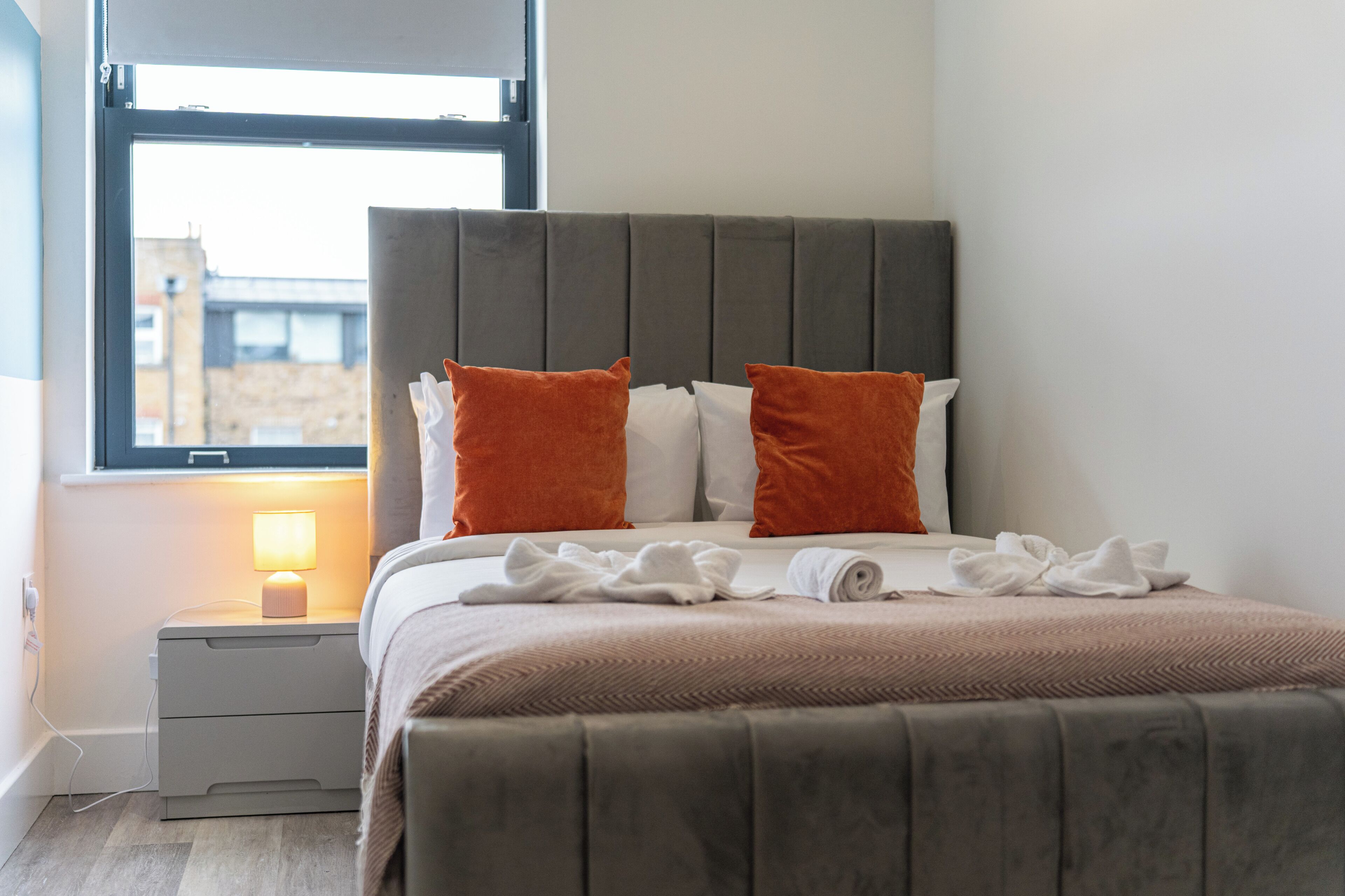 Ein ordentlich gemachtes Bett mit Kopfteil, ein Nachttisch mit Lampe und ein Fenster mit Blick auf Gebäude.