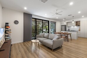 Ein modernes Wohnzimmer mit einer Couch, einem kleinen runden Tisch und einem Essbereich mit Stühlen. Es gibt eine Küchenzeile mit Kühlschrank und Spüle.
