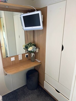 Hotellihuoneessa on puinen peilipöytä, peili, pieni roskakori, valkoinen vaatekaappi ja seinälle asennettu televisio.