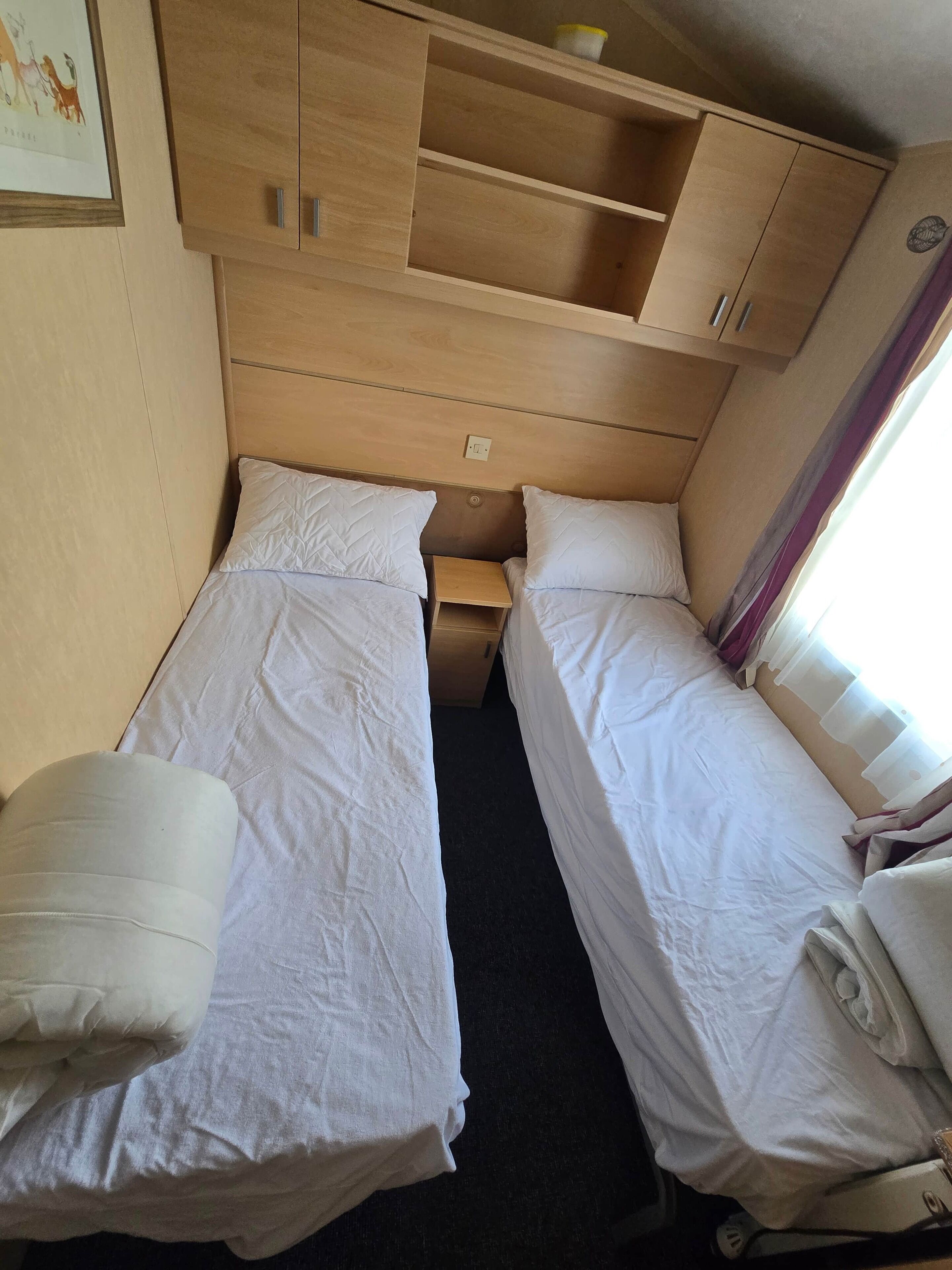 A compact room with two single beds, a small wooden shelf unit, and a power outlet on the wall.