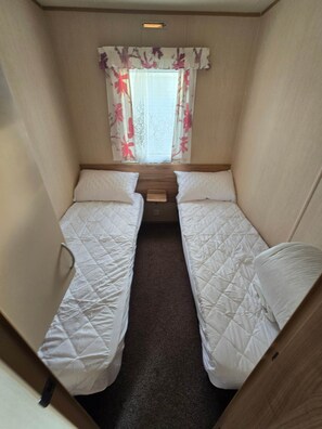 A narrow room with two beds, a window with floral curtains, and a small wooden table. - Towyn Oakfields  F70 (Kinmel Bay)