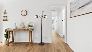 A hallway with a wooden floor, a standing lamp, a wall clock, a framed painting, and a potted plant. - Oceanview 3-Bedroom Apartment in Kiama (Kiama)