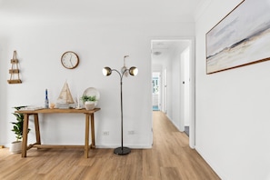 A hallway with a wooden floor, a standing lamp, a wall clock, a framed painting, and a potted plant. - Oceanview 3-Bedroom Apartment in Kiama (Kiama)
