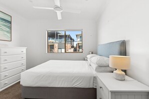 Ein Schlafzimmer mit Bett, Nachttisch, Lampe, Kommode, Deckenventilator und Fenster mit Blick auf die Stadt.