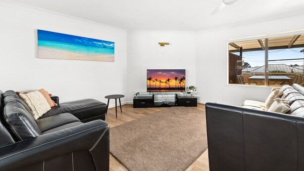 A living room with a black leather sofa, a glass coffee table, a flat-screen TV, a ceiling fan, and a large window with a view of a patio. - Oceanview 3-Bedroom Apartment in Kiama (Kiama)