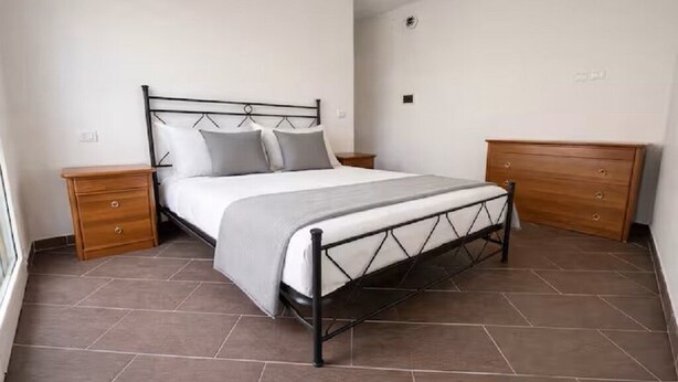 Quarto com estrutura de cama de metal, mesa de cabeceira de madeira, cômoda de madeira e piso ladrilhado.