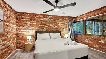 A room with a brick wall, a bed with white bedding, a ceiling fan, and a window with curtains.
