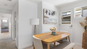 Un coin repas avec une table en bois, des chaises rembourrées, un vase de fleurs et un tableau sur le thème de la plage accroché au mur.