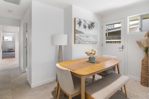 A dining area with a wooden table, cushioned chairs, a vase with flowers, and a beach-themed painting on the wall. - Spacious 4-Bed Coastal Home with Pool (Mooloolaba)