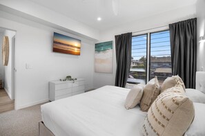 A modern bedroom with a large bed, a nightstand, a dresser, and a window with curtains. - Spacious 4-Bed Coastal Home with Pool (Mooloolaba)