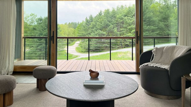 A room with a large sliding glass door, a round table with a book and a bowl, a grey armchair, and a wooden ceiling.