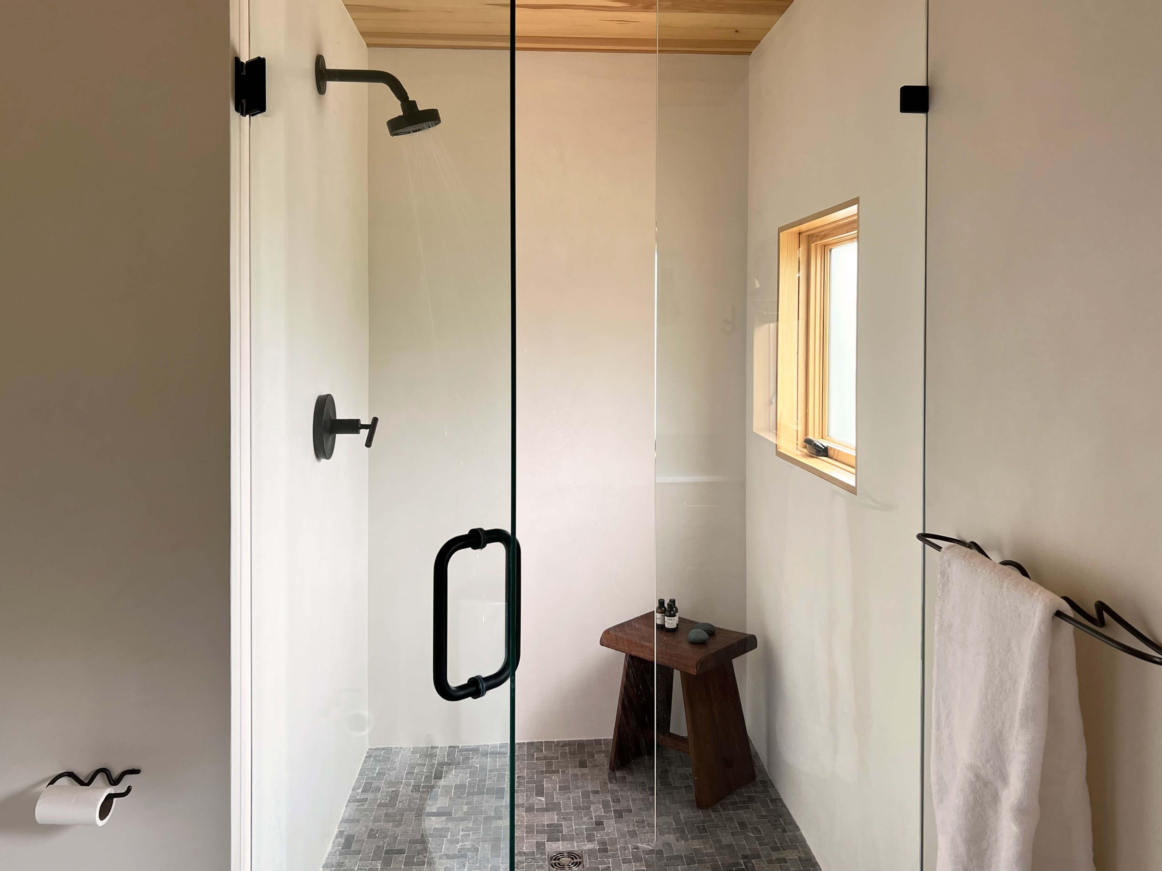 A modern bathroom with a glass shower enclosure, a small wooden stool, and a window providing natural light.
