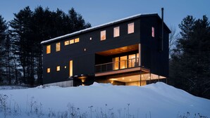 A modern two-story house with a balcony, large windows, and a snowy landscape.