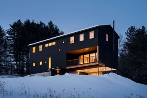 Una casa moderna de dos pisos amb un balcó, finestres grans i un paisatge cobert de neu.
