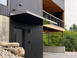 Un edifici modern amb una façana vertical destacada, un balcó amb barana de fusta i una estructura de pedra al primer pla.